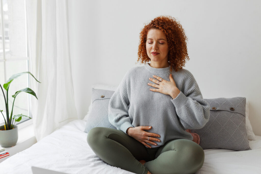 A female in casual clothes performing mindfulness meditation with her eyes closed while seated on a bed next to window.